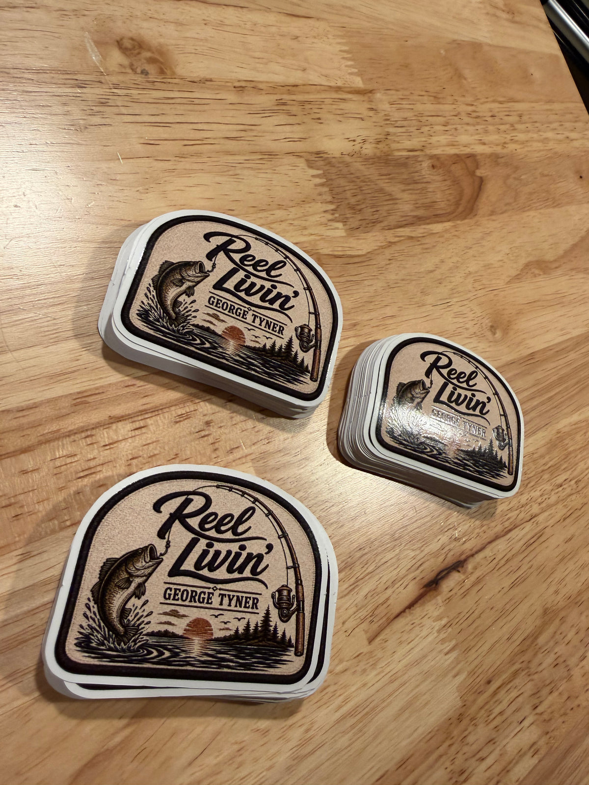 Three stacks of Reel Livin' fishing stickers printed on white vinyl, on a wooden table.