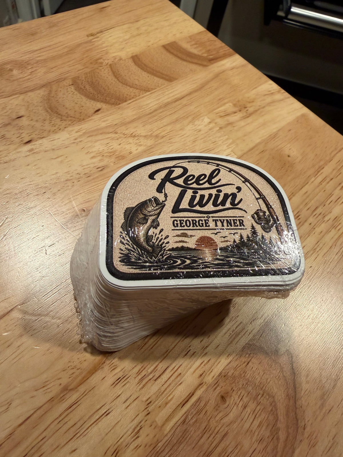 Stack of Reel Livin' fishing stickers with a bass, fishing rod and sunset design, wrapped on a wooden table.
