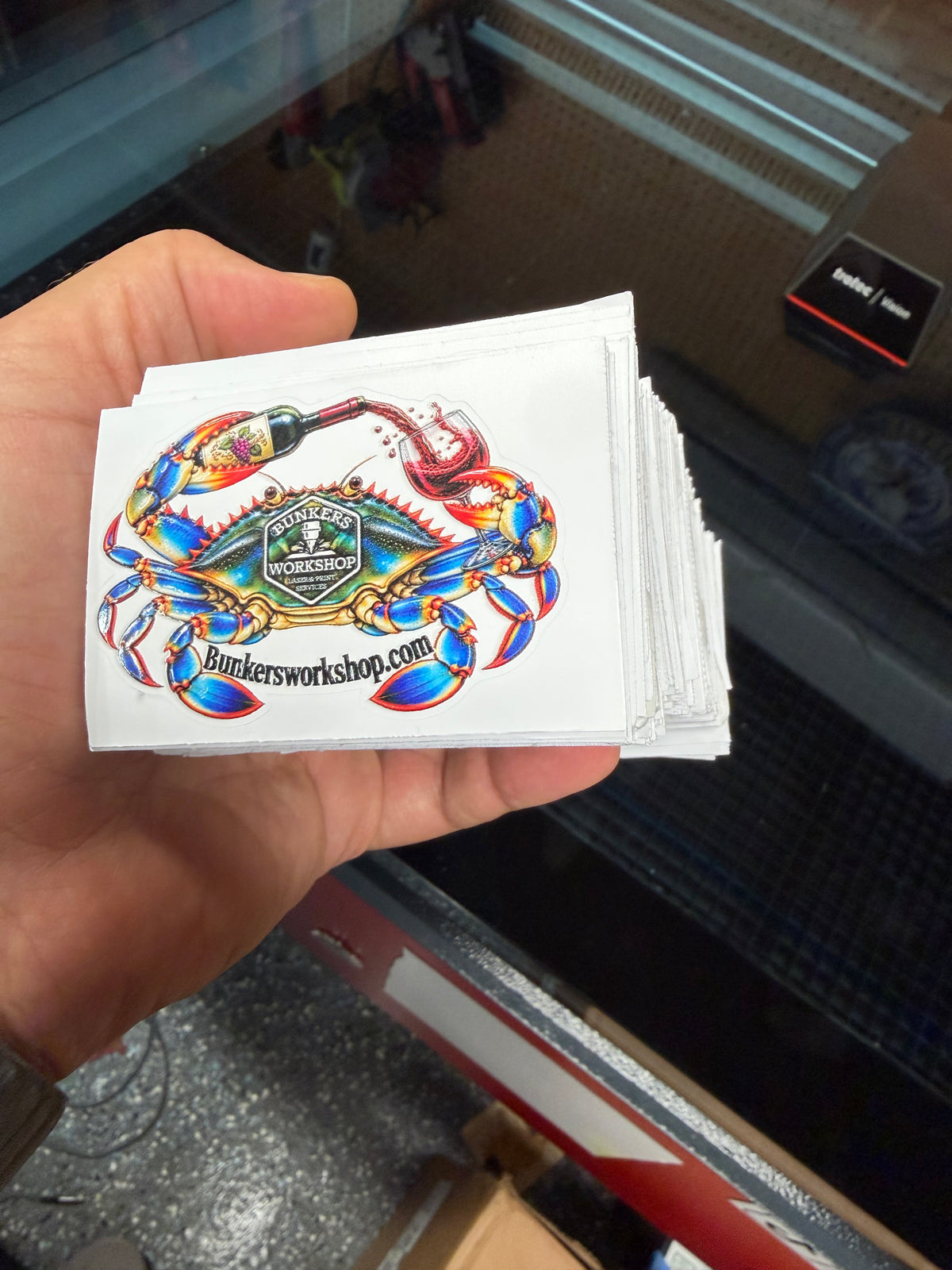 Hand holding a stack of Bunkers Workshop crab logo stickers with a wine bottle and glass.