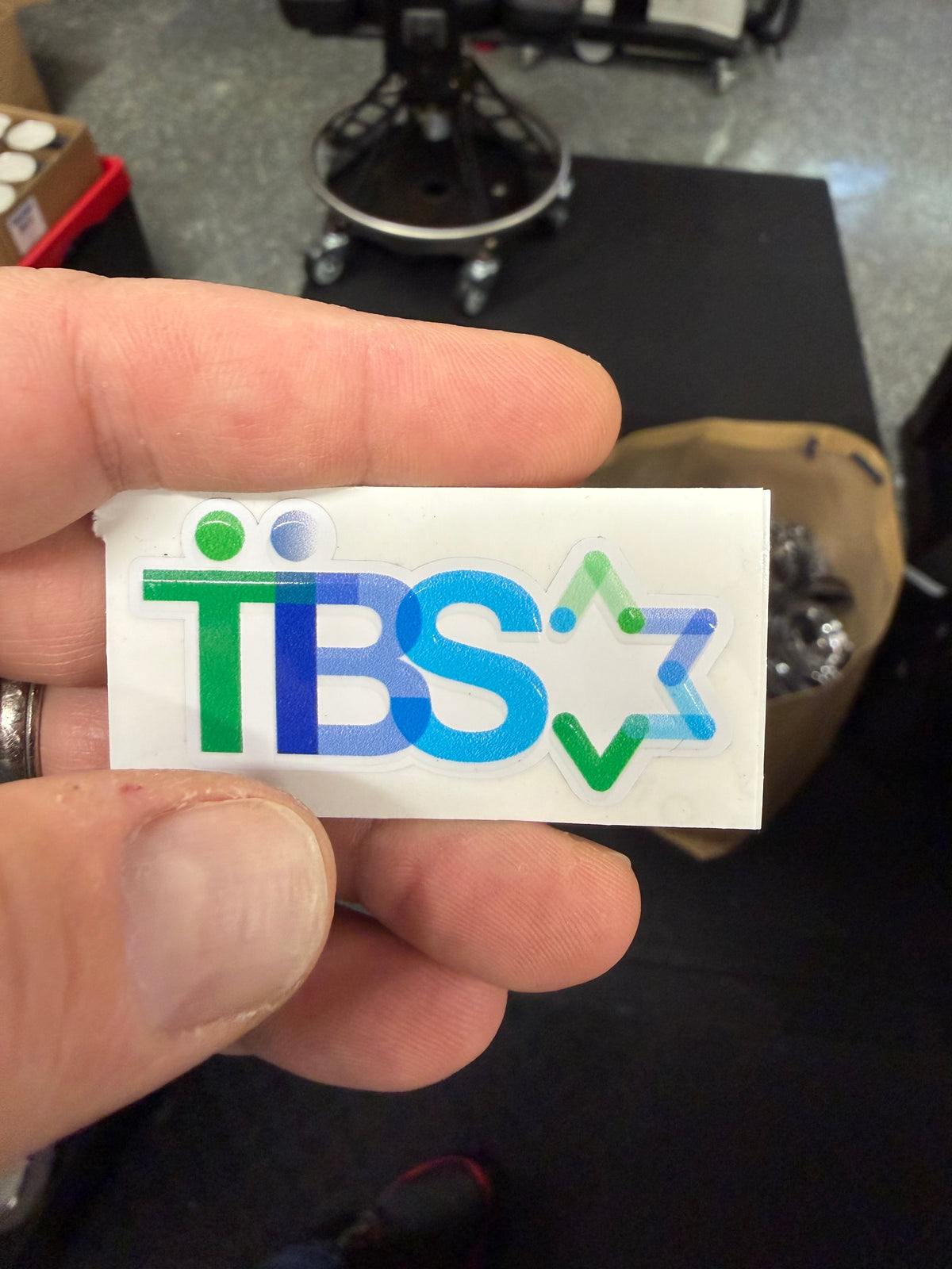 Hand holding a blue and green TBS logo sticker with a six-pointed star on white backing.