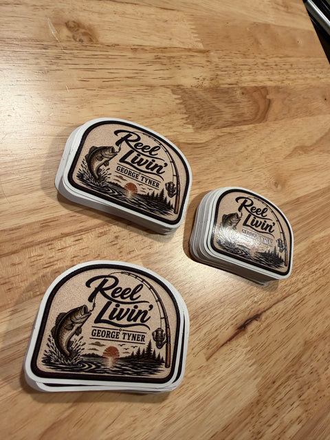 Three stacks of Reel Livin' fishing stickers printed on white vinyl, on a wooden table.