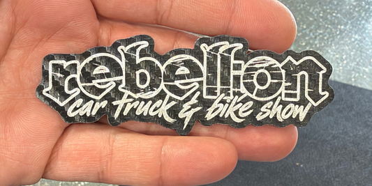 Close-up of a black Rebellion logo laser-engraved patch held in a hand, showing the complete engraved design.